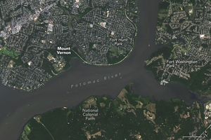 Image centers the Potomac River running primarily horizontal across the image. Below the river the land is mostly green including open fields and the National Colonial Farm. Fort Washington Park and sprawling suburbs are seen at the edge of a wide fork in the river.  Above the River is more green land labeled Mount Vernon and Fort Hunt Park to the right. The land around this area shows signs of significant human settlement.