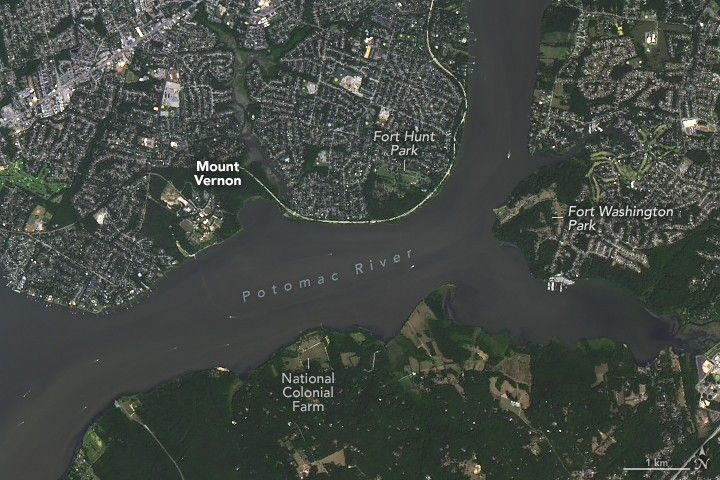 Image centers the Potomac River running primarily horizontal across the image. Below the river the land is mostly green including open fields and the National Colonial Farm. Fort Washington Park and sprawling suburbs are seen at the edge of a wide fork in the river. Above the River is more green land labeled Mount Vernon and Fort Hunt Park to the right. The land around this area shows signs of significant human settlement.