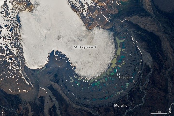 The Múlajökull glacier appears white and dominates the image flowing from the upper right to toward the lower right. There are other snow filled valleys in the upper left and the lower right is filled with reflective puddles between labeled Drumlins and in an arc beyond the edge of the glacier is the Moraine, where rock had previously been pushed by the glacier. Streams meander away from the glacier in multiple directions.