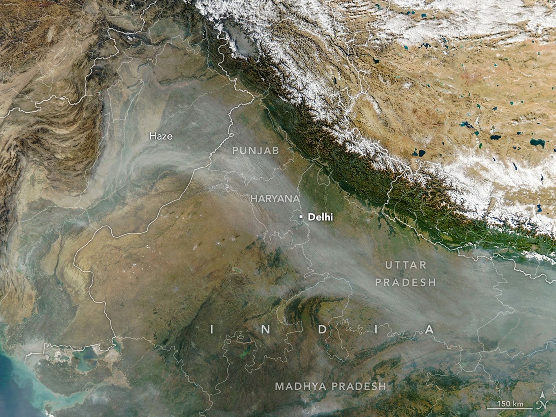 A satellite image of northern India shows a plume of smoke and haze spreading across the Indo-Gangetic Plain. Gray haze is visible over the Indian states of Punjab, Haryana, Uttar Pradesh, and Madhya Pradesh, which are labeled. The snow-covered mountains of the Tibetan Plateau are visible to the north.