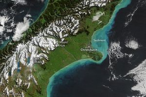 a long fairly narrow island, with water on either side stretches diagonally from bottom left to upper right. Off the shore to the right of the island the waters are turquoise, labeled as sediment. The city of Christchurch resides just right of center on the coastline. Mount Hutt is directly left of Christchurch in the white snow covered mountains. The land between and near the right coastline is largely green in color. In the lower left Lake Tekapo is labeled among the snow covered mountains.