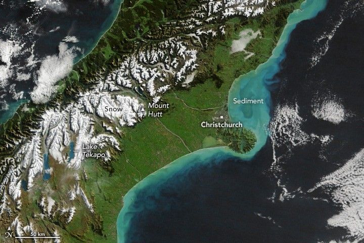 a long fairly narrow island, with water on either side stretches diagonally from bottom left to upper right. Off the shore to the right of the island the waters are turquoise, labeled as sediment. The city of Christchurch resides just right of center on the coastline. Mount Hutt is directly left of Christchurch in the white snow covered mountains. The land between and near the right coastline is largely green in color. In the lower left Lake Tekapo is labeled among the snow covered mountains.