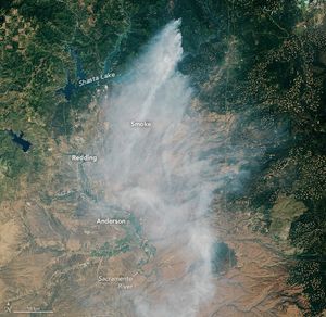Wildfire smoke emanates from the top of the image where the land is covered in more foliage, just right of Shasta Lake.  The smoke spreads southward toward Redding, Anderson and the lower Sacramento River which have a more arid brown landscape.