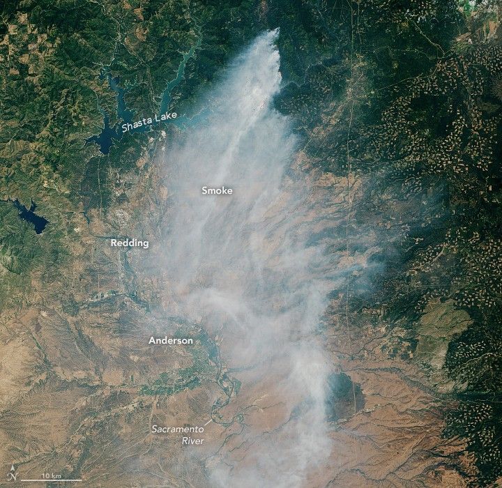 Wildfire smoke emanates from the top of the image where the land is covered in more foliage, just right of Shasta Lake. The smoke spreads southward toward Redding, Anderson and the lower Sacramento River which have a more arid brown landscape.