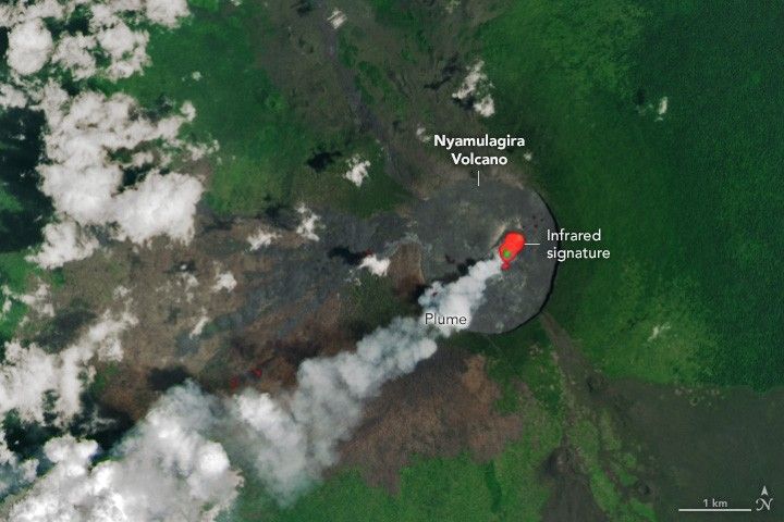 A bright red infrared signature is nestled in a matte black disk labeled as Nyamulagira Volcano. Smoke emanates from the infrared signature and drifts to the lower left corned of the image and is labeled Plume. The volcanic disk appears sunken compared to the surrounding land which is a rich green color. to the lower left the land is more brown, likely due to volcanic activity, and partial cloud cover obscures the leftmost side of the image.
