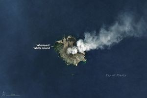 Whakaari is shown in the medium blue waters of the Bay of Plenty. The island is emitting whitish smoke, which is drifting to the right side of the image. The island is mostly bare and gray with some green foliage on the slopes of the upper end of the island.