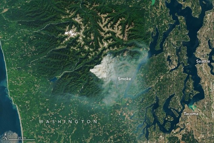 Smoke emanates from the deep green foliage covering the craggy landscape left of Seattle. Tacoma is also visible in the lower right corner of the image.
