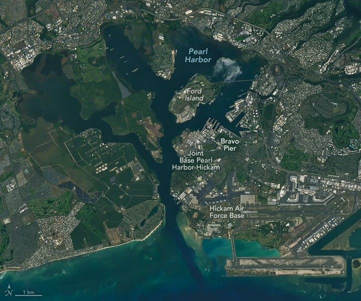 The image features Pearl Harbor, an expansive inland coastline with peninsulas and Ford island within it, resides behind the coastal inlet. Near the image center, just below Ford Island, is Join Base Pearl Harbor-Hickam, to the right of which is Bravo Pier. This section of the imaged area is densely built up with white top buildings. in the lower right portion of the image a long stretch of Tarmac appears to be the runway of Hickam Air Force Base. The image is filled with densely built home and business structures, with sporadic patches of deep green foliage.