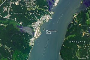 Image of the Potomac River at Quantico. The Chopawamsic island is within the river closer to the Virginia side near the center. The creek by the same name is visible behind the concrete structures of Quantico. The left and right sides of the image are majority covered in deep green foliage with Virginia being on the left and Maryland on the right.