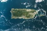 From above, a rectangular green and brown island sits amid blue waters. The main island is fringed by smaller islets and dotted with wisps of white clouds.