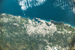A detailed view of the island’s north side shows tan urban areas along the coast and inland, surrounded by green vegetation. The upper third of the image features blue ocean waters dotted with white clouds.