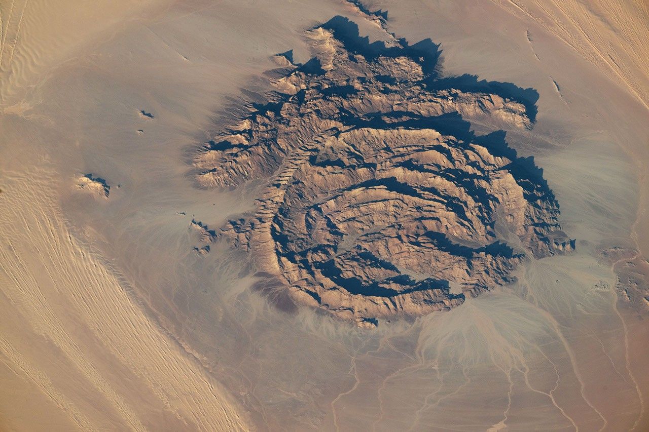 A top-down view shows circular rocky formations rising from a flat, sandy-brown landscape. Darker brown sand encircles the rocky rings, with lighter patches of outwash spreading across the terrain.