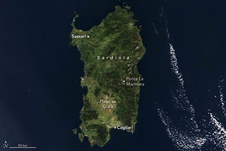 The green island of Sardinia centers in the image, a few wisps of clouds in the lower right, but otherwise surrounded by navy blue seas. Sassari is labeled in the upper left corner of the island that appears roughly twice as tall as wide. Along the right center of the island is Puna La Marmora. The lower coast is home to Cagliari which is the coastal end of a valley that stretches up and right. The extent of the reach up is Planu sa Giara, where the land is a deep green color with foliage.