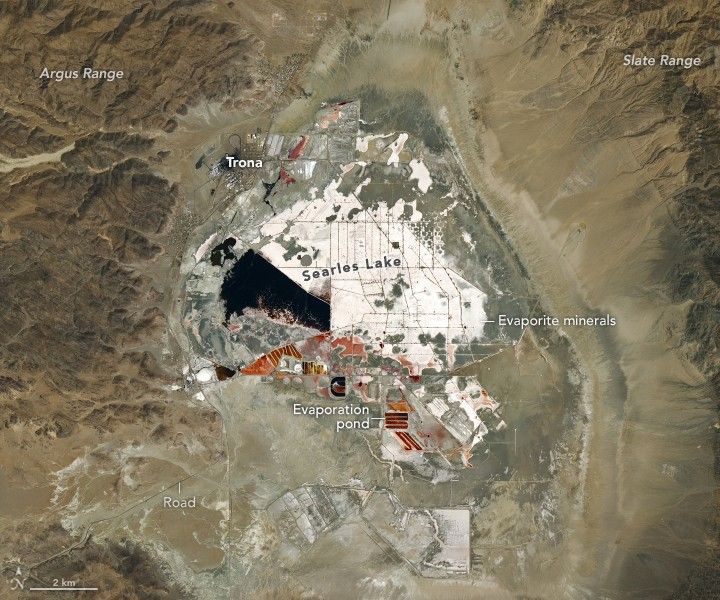 In a valley between the Argus Range, left and the Slate Range, right, is the Searles Lake. The lake is partitioned into numerous sections where Evaporation ponds, seen along the bottom of the lakebed region leave evaporite minerals, white in color, in sectioned off lake fields. Actual water body is seen on the left side of the Searles Lake, below the labeled settlement of Trona. A road leads in to this region from the bottom left.
