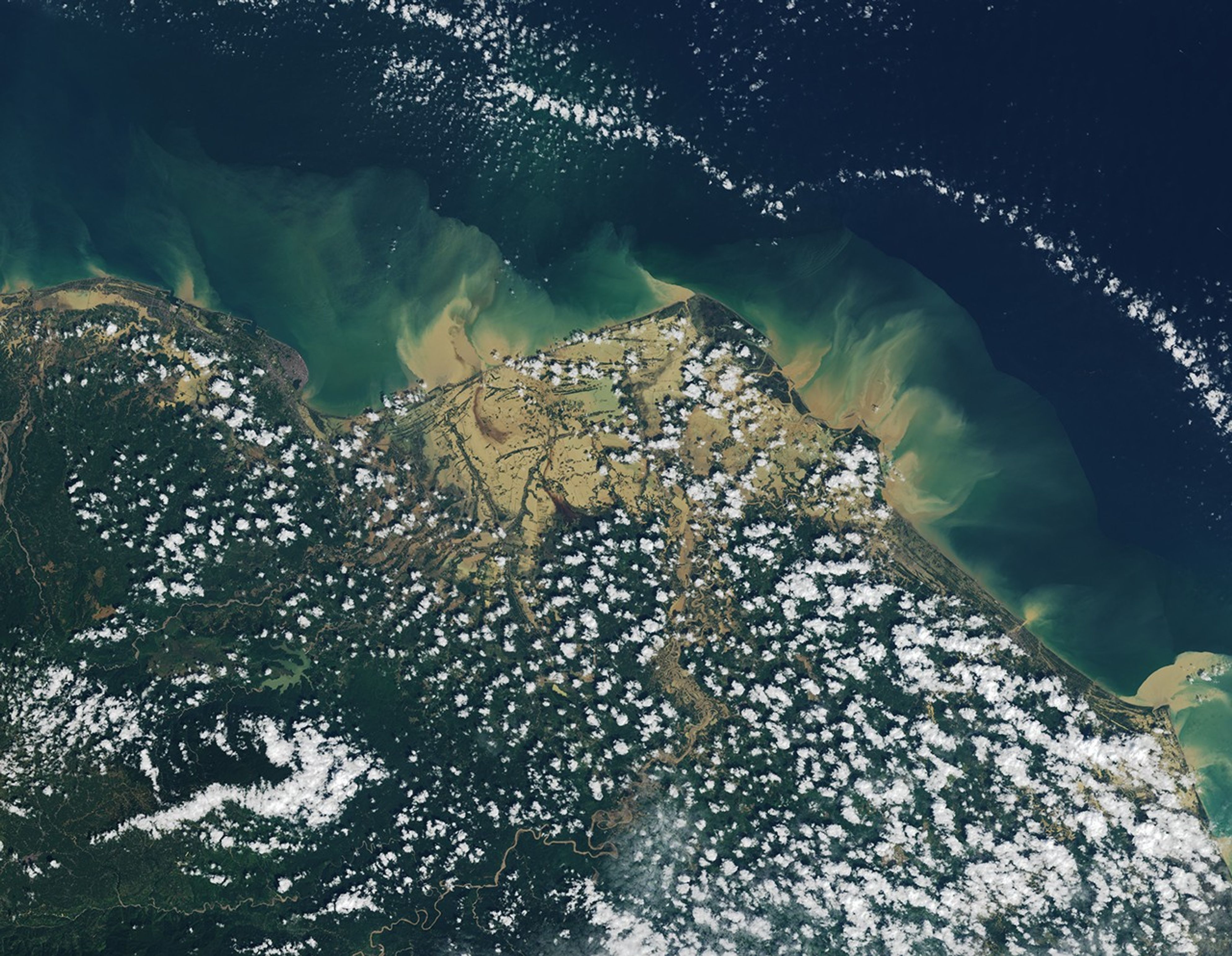 A satellite image centered on the town of Lhoksukon in Sumatra shows brown, muddy floodwater covering much of a coastal plain near the town. Plumes of brown water are visible mixing with the bluer waters of the Strait of Malacca in the upper part of the image. The lower part of the image is greener, more mountainous, and speckled with small clouds.