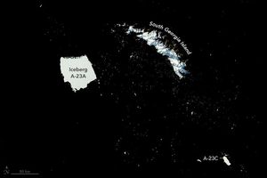 The deep near black sea is seen dotted with white specs and three major white structures. South Georgia island is an arced  snow covered and craggy landscape. To the left of there is Iceberg A-23A, which is mostly square in shape and on a diagonal orientation to the image. It also occupies about as much space in the image as South Georgia Island. A-23C is much smaller and in the lower right.
