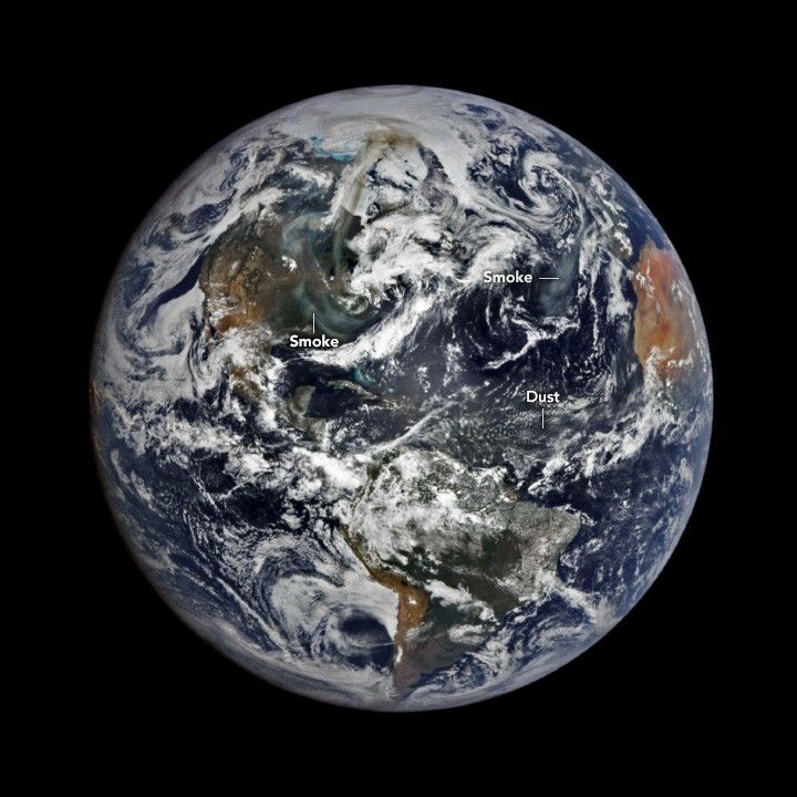 Image of the Earth zoomed out to span the entire Americas and part of Africa. The visible land is primarily green to dusky brown in color, aside from Northern Africa, which is a bright tan color. Clouds are interspersed across the entire image obscuring much of the land and blue ocean below, but a slight discoloration between Africa and South America is labeled as dust. A significant darkened swirl coupled against the swirling clouds over North America. This is labeled as smoke.