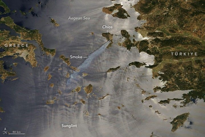 The island of Chios, between Greece, left, and Türkiye, right, is in the upper center of the image in the gray-blue aegean sea.  Smoke streams from Chios down and left from the island.