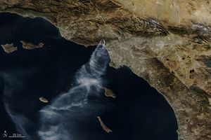 Southern California coastline centers Los Angeles and the Pacific Ocean. The Pacific fills the lower left half of the image with the coastline running diagonal. To the left of central Los Angeles is the Palisades fires. Smoke emanating from those hills streams out over and disperses over the Ocean. Above the Los Angeles label in this image is the Eaton fire. This more inland fire is generating less smoke than the palisades, but it also disperses toward the coast.