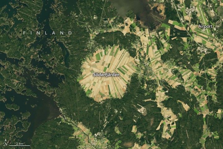 In Finland, a circular region of land is completely divided into crop fields and labelled Söderfjärden. An airport is in the upper right corner. To the left of Söderfjärden the land is broken up by waterways and small islands covered in dense dark green foliage.  There are more fields of crops between Söderfjärden  and the airport that seem to follow valley's leaving higher elevations covered in dark green foliage. The fields are all a light green to beige color.