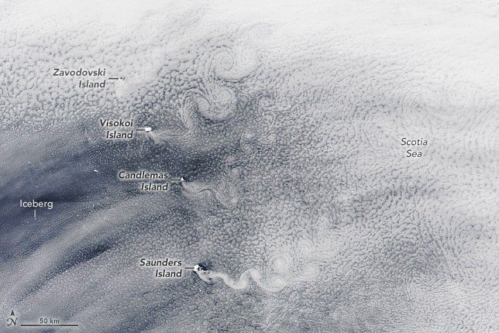 Swirling white clouds blanket the image with thin edges giving a glimpse of the blue waters below. The right side of the image is labeled Scotia Sea. The middle of the left labels an iceberg in the region that has the thinnest cloud layer. A line of  islands spanning the image vertically is the point at which clouds begin to swirl as the density increases to the right.
