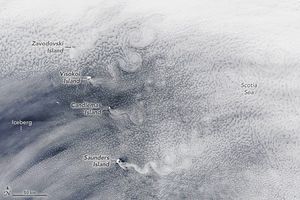 Swirling white clouds blanket the image with thin edges giving a glimpse of the blue waters below. The right side of the image is labeled Scotia Sea. The middle of the left labels an iceberg in the region that has the thinnest cloud layer. A line of  islands spanning the image vertically is the point at which clouds begin to swirl as the density increases to the right.