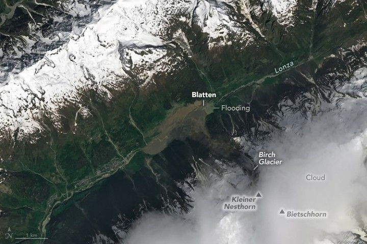 Snow covered mountain ridge occupies the upper left  to top center of the image. They Valley below contains the river Lonza. In the center of the image Blatten is labeled along the river along with flooding. The area is covered in a medium brown amorphous slurry. Another ridge is hidden by clouds in the lower right and includes the Birch Glacier, Kleiner Nesthorn Mountain, and Bietschhorn.