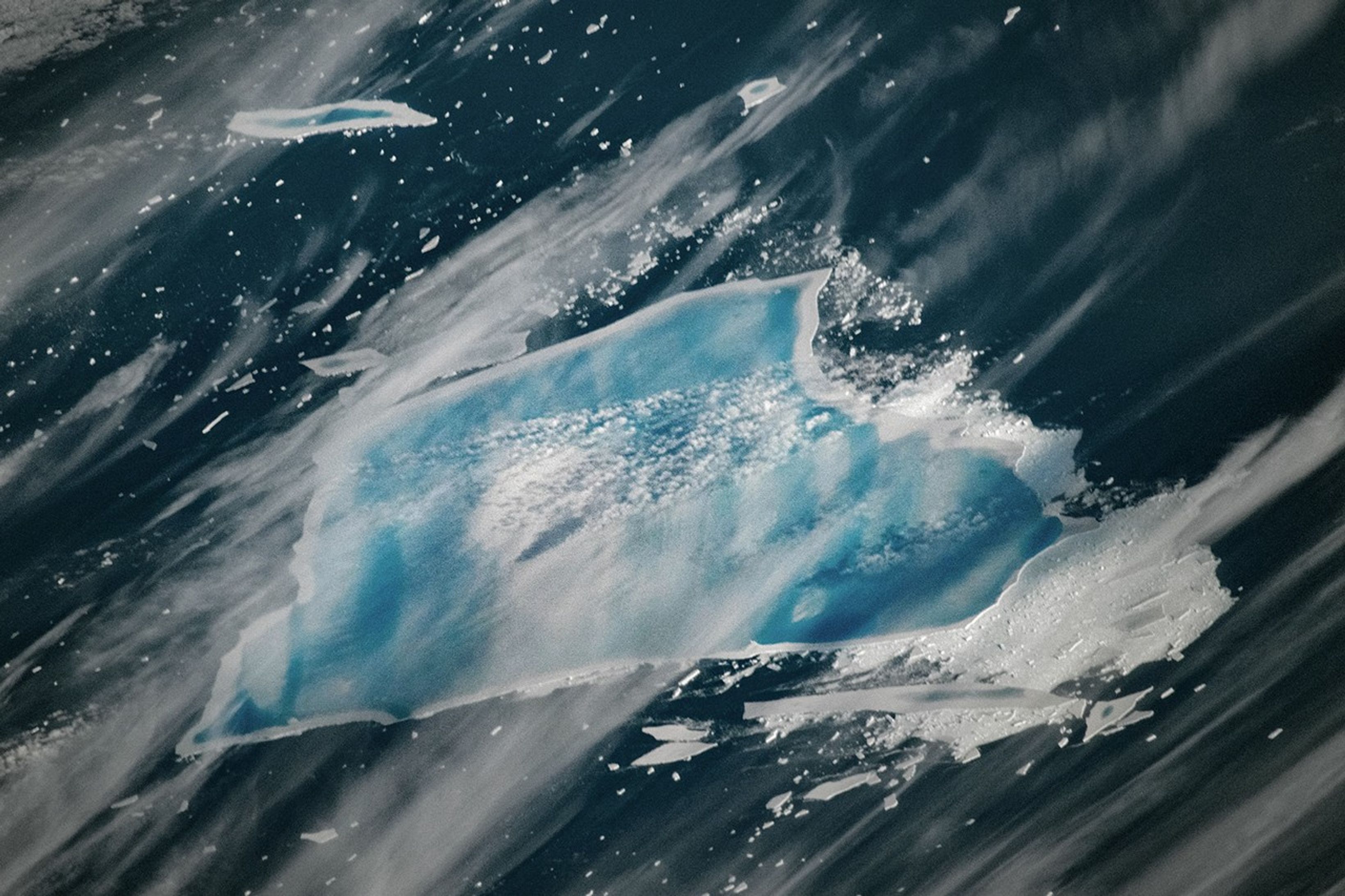 A photograph taken by an astronaut aboard the International Space Station on December 27, 2025, shows a closer view of the iceberg. Striking blue melt ponds cover nearly the surface top of A-23A except for a thin boundary of white along the edge—an area where the ice has warped upward. Wispy clouds run diagonally across the image, partially obscuring the view.