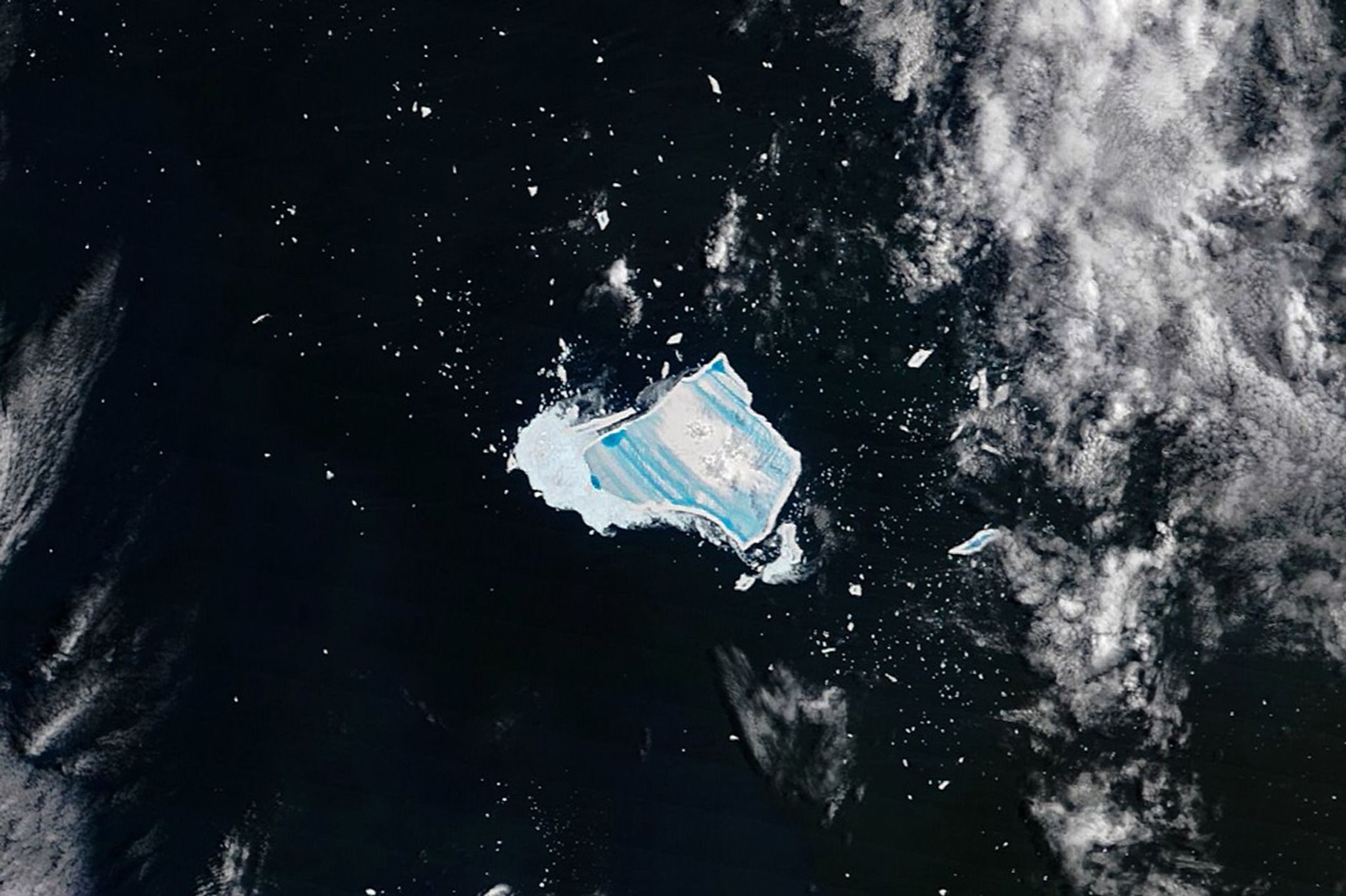 A satellite image captured on December 26, 2025, centers on iceberg A-23A adrift in dark Southern Ocean waters. The iceberg appears as a bright white, roughly rectangular slab with jagged edges and parallel blue stripes across its surface. The blue areas are meltwater ponds. A thick patch of ice fragments known as brash ice is visible along one edge of the main iceberg in an area where meltwater is leaking from it, while patches of small icebergs and clouds are also visible around it.