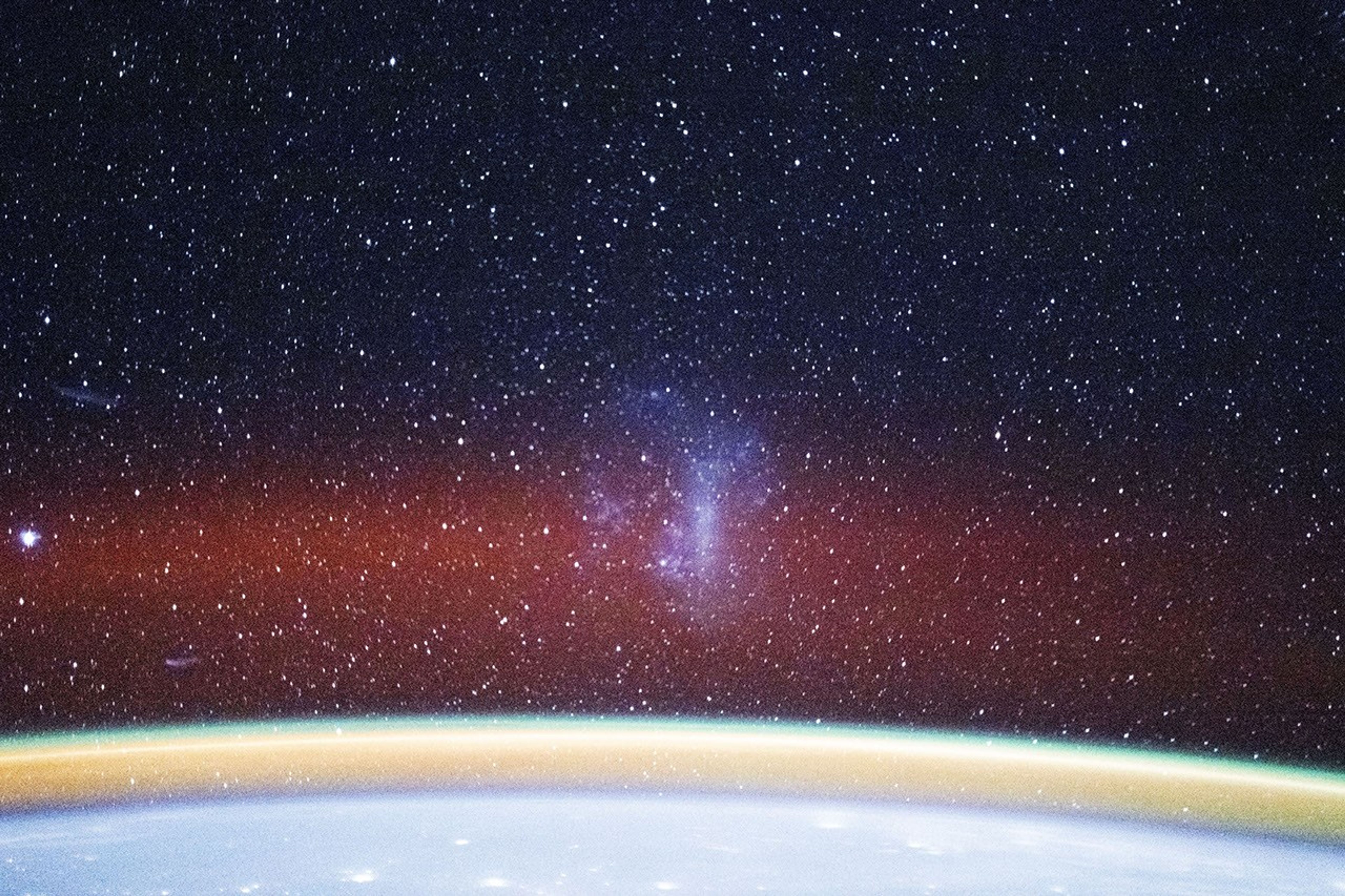 A photo taken from the International Space Station looks out over Earth’s horizon into space. Many white points of light appear against a dark background. A bright blurry area near the center of the photo is the Large Magellanic Cloud, a nearby dwarf galaxy. Earth’s bright blue horizon and layers of bright yellow and faint orange airglow arc across the bottom of the image.