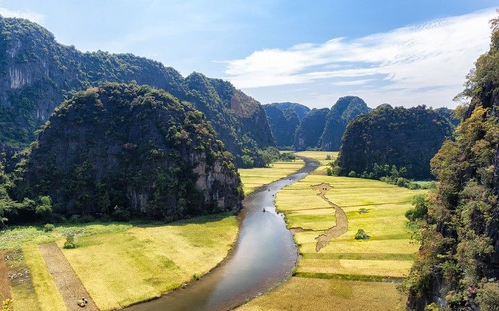 This photograph looks out over a valley where a calm river meanders through yellow fields. Steep-sided rock towers covered in green vegetation rise from both sides of the valley.