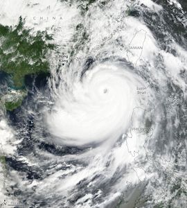 Swirling white clouds off the coast of China, also mostly covered by clouds with some green foliage covered landscape visible. Taiwan and the Philippines are both completely obscured by clouds, but the island outlines are overlaid on the image.