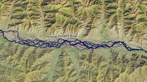 The Yarlung Zangbo River braids across the valley that runs horizontally across this image. Mountainous peaks fill the landscape both above and below the river..