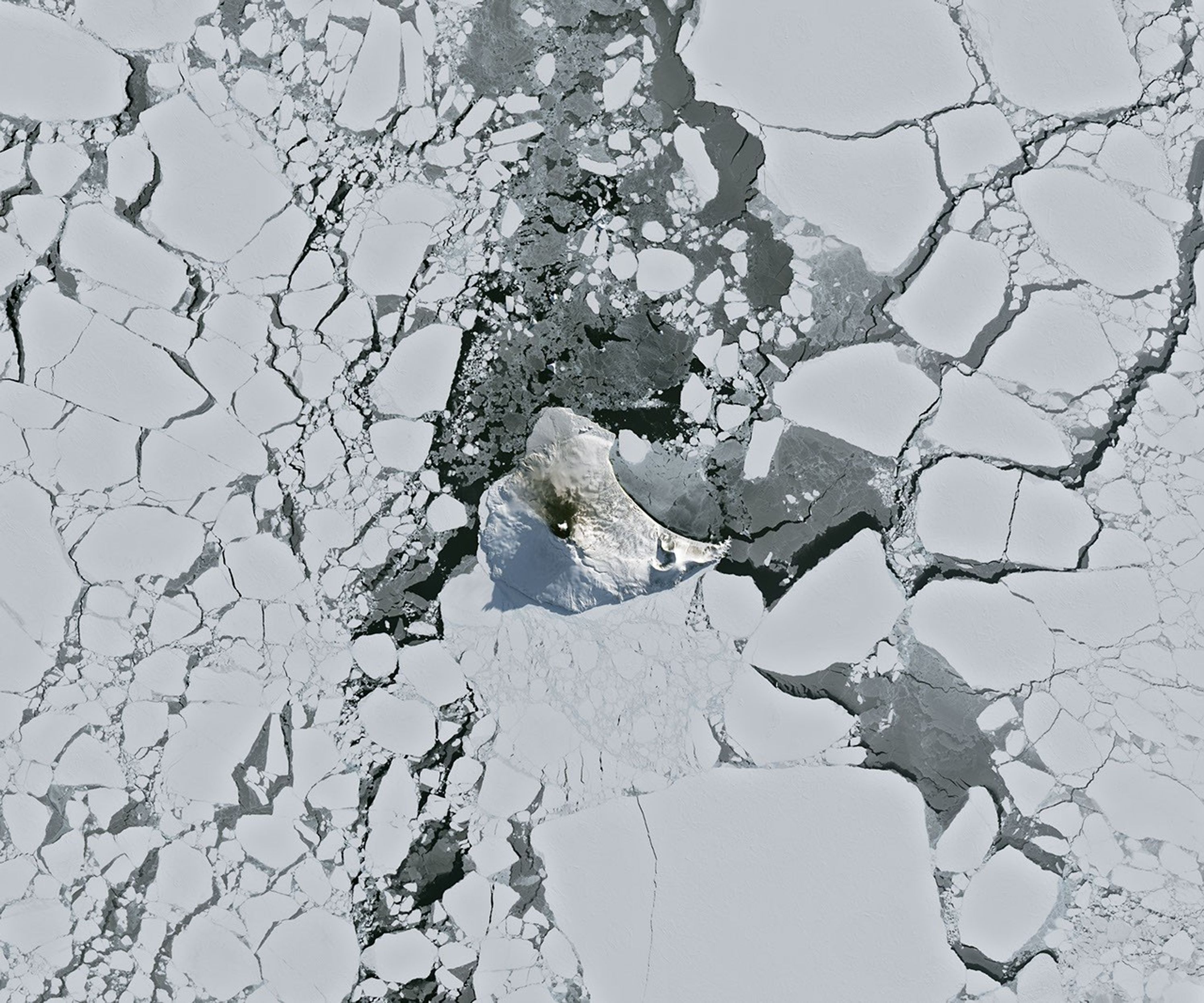 A small ice- and snow-covered island with an active volcanic crater at its center is surrounded by drifting pieces of sea ice. A thermal signal and small plume appear in the crater, and ash darkens the snow on the volcano’s northern slopes.