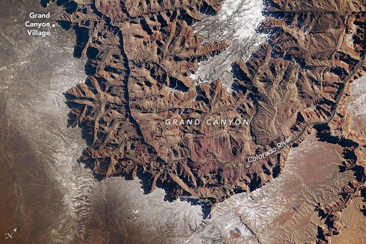 A portion of the Grand Canyon, photographed from above, forms a U shape. The plateau on both sides of the canyon is partially covered in a layer of white snow.