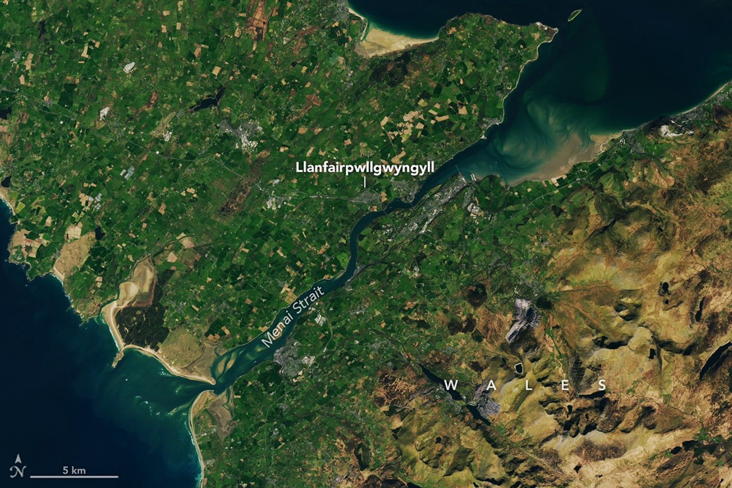 A wider view of the same area shows how the Strait of Menai connects to the open ocean to the north and south of Anglesey island. The right side of the image transitions to hillier terrain with less farmland.