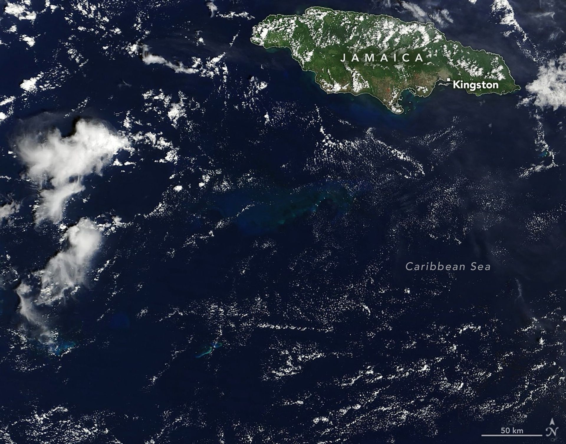 A satellite image shows a portion of the dark blue Caribbean Sea near Jamaica. A submerged carbonate platform appears as a slightly brighter blue area of water in the center. The mostly green island of Jamaica is in the upper right, and scattered clouds are present throughout.