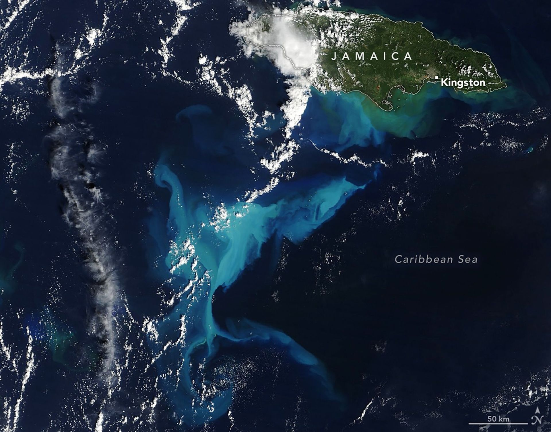 A satellite image shows a portion of the Caribbean Sea near Jamaica. Much of the water in the middle third of the image is bright blue due to suspended sediment. The mostly green island of Jamaica is in the upper right, and scattered clouds are present throughout.