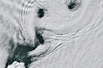 Parallel lines of cloud vortices appear downwind of a small, ice-covered island.