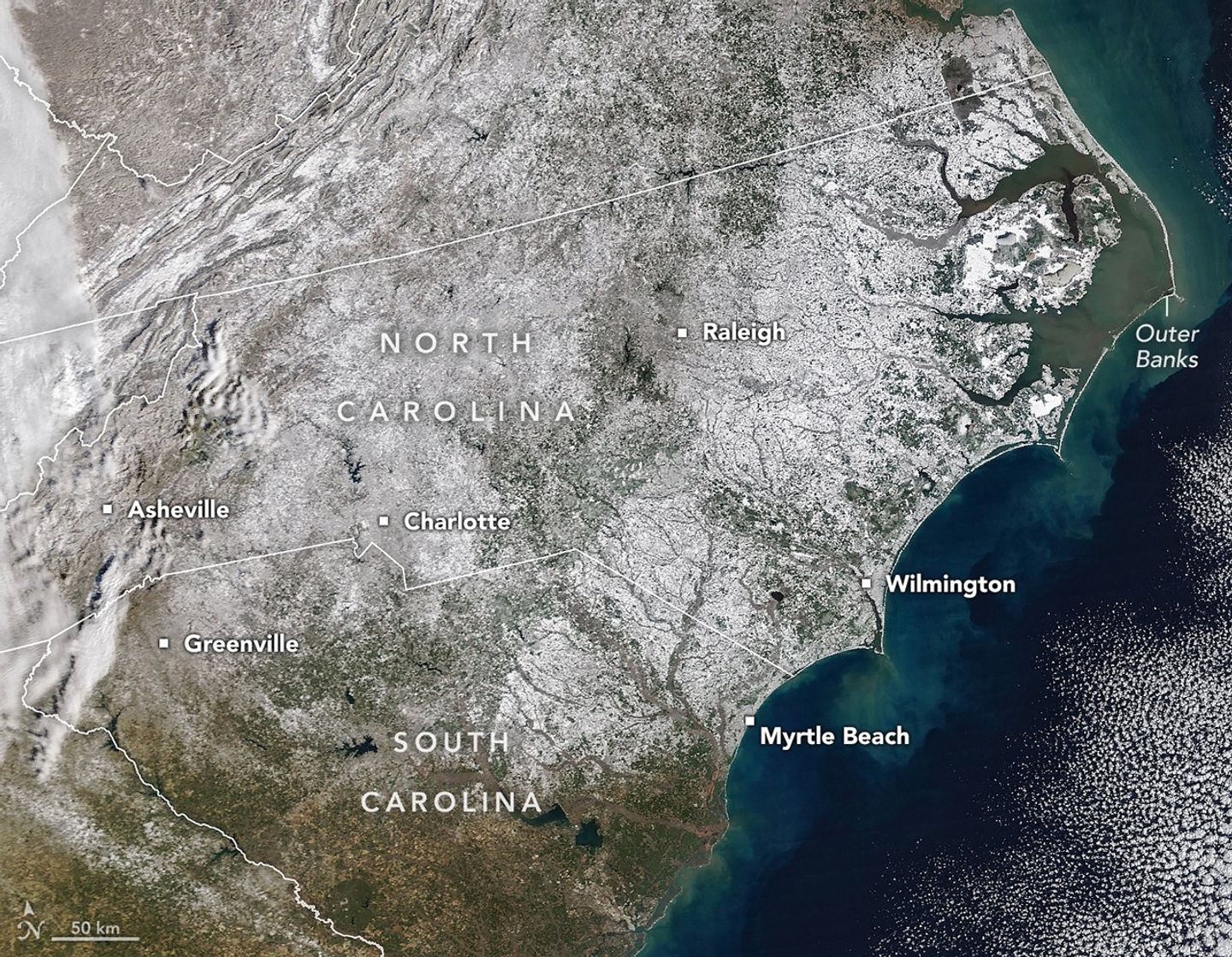 A satellite image centered on North Carolina shows white snow covering most of the state, with additional snowy patches in neighboring states. Some forested and urban areas appear gray. To the right, a strip of barrier islands appears white against the green and blue Atlantic Ocean.