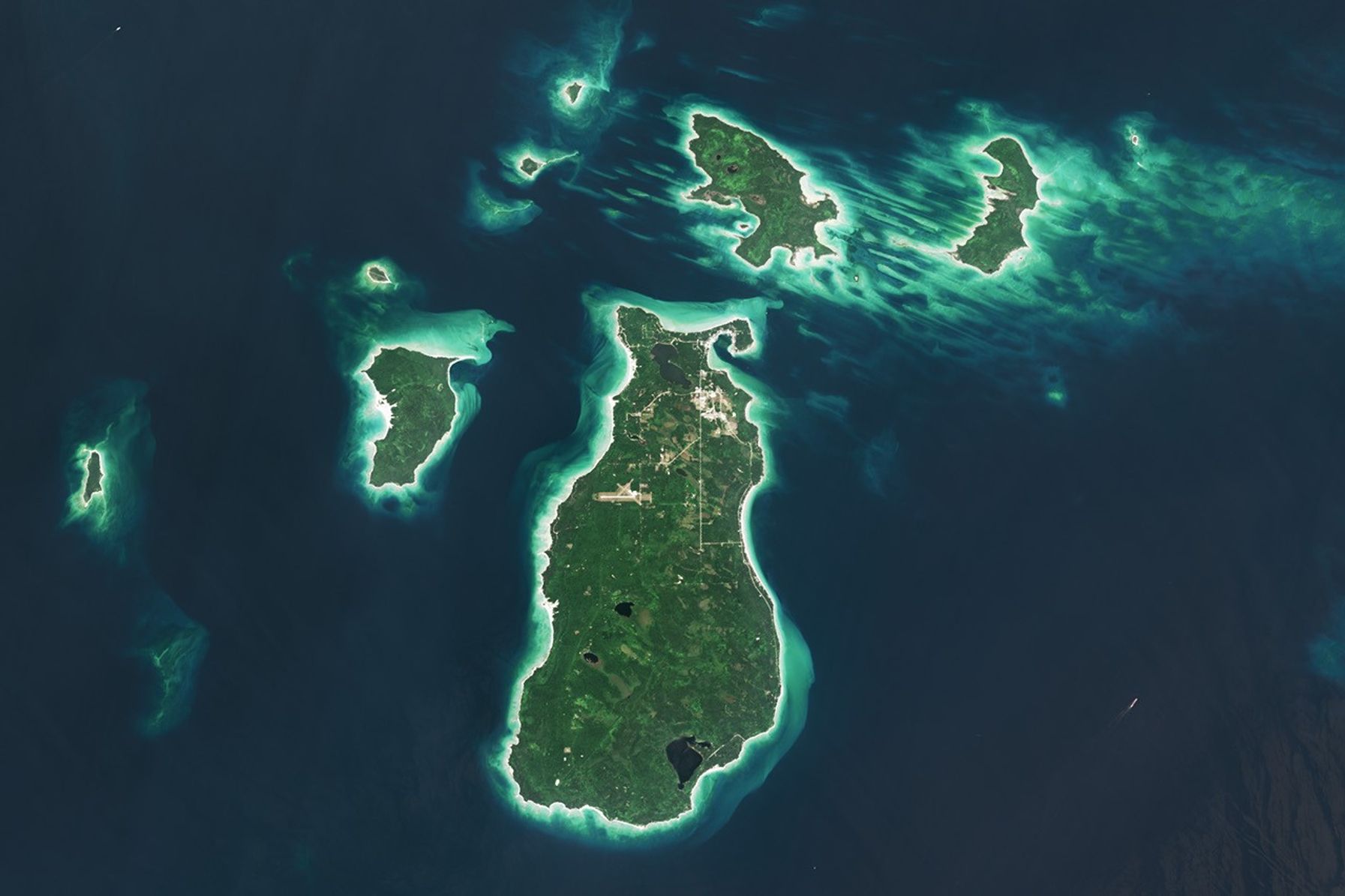 A satellite image shows several islands in Lake Michigan. The largest one, Beaver Island, is in the center. The islands are mostly green and vegetated, with bright sandy areas on their perimeters. Shallow waters near the land appear turquoise, and deeper waters are dark blue.