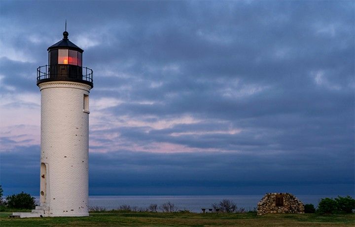 A lighthouse with a white tower and a glowing red beacon at the top stands on the left side of this photograph. A large lake and cloudy skies fill the background.