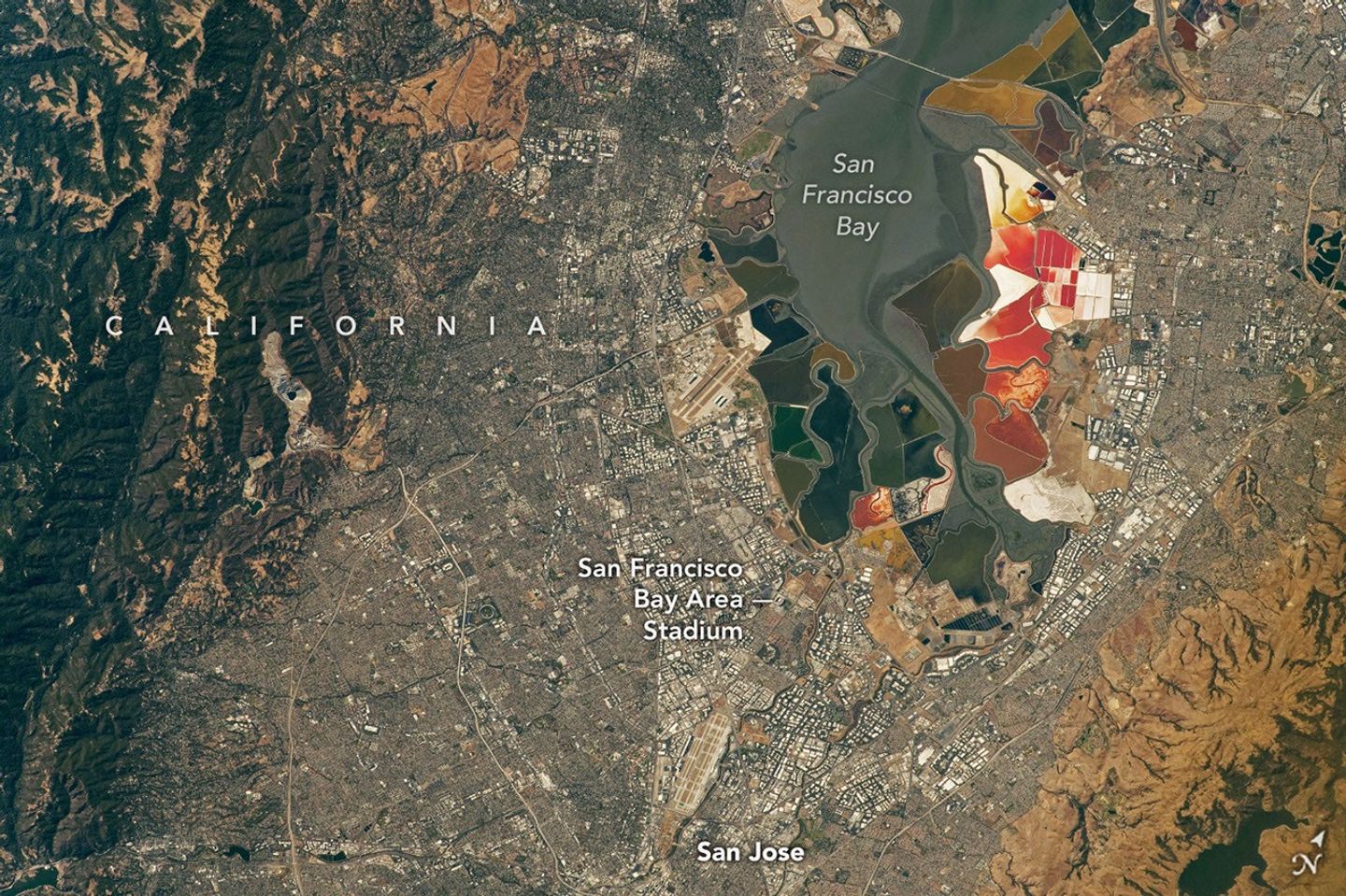 An aerial view of the San Francisco Bay Area Stadium, middle-left, is surrounded by urban infrastructure. Colorful ponds of the southern San Francisco Bay are visible toward the top.