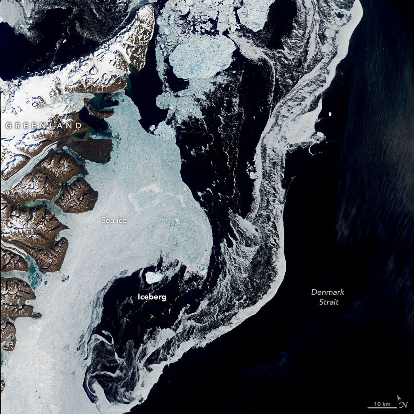 A satellite image shows the dark blue waters of the Denmark Strait with swirling sea ice to the right of eastern Greenland. A bright white iceberg sits amid the sea ice in the middle-left of the scene.