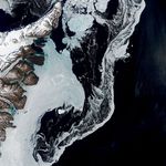 A satellite image shows the dark blue waters of the Denmark Strait with swirling sea ice to the right of eastern Greenland. A bright white iceberg sits amid the sea ice in the middle-left of the scene.