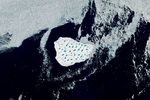 A detailed view shows the iceberg's bright white surface pocked with light blue meltwater ponds. Broken pieces of sea ice float around the iceberg in otherwise dark blue ocean water.