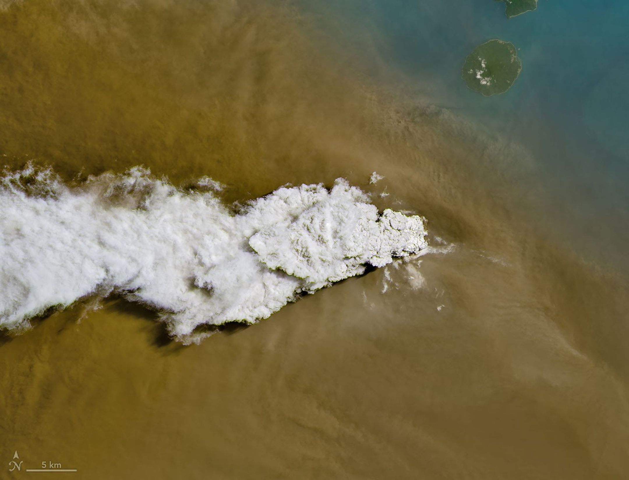 A white volcanic plume streams left over a tan ash cloud that fills most of the image. A small green island and blue water are visible in the upper right.