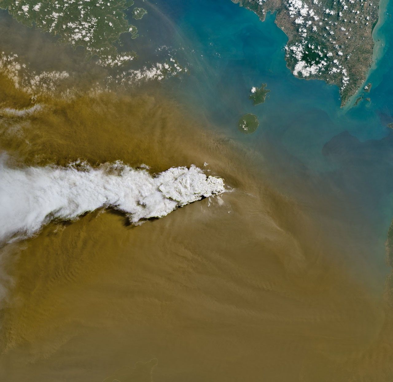 A white volcanic plume streams left over a tan ash cloud that fills most of the image. A small green island and blue water are visible in the upper right.