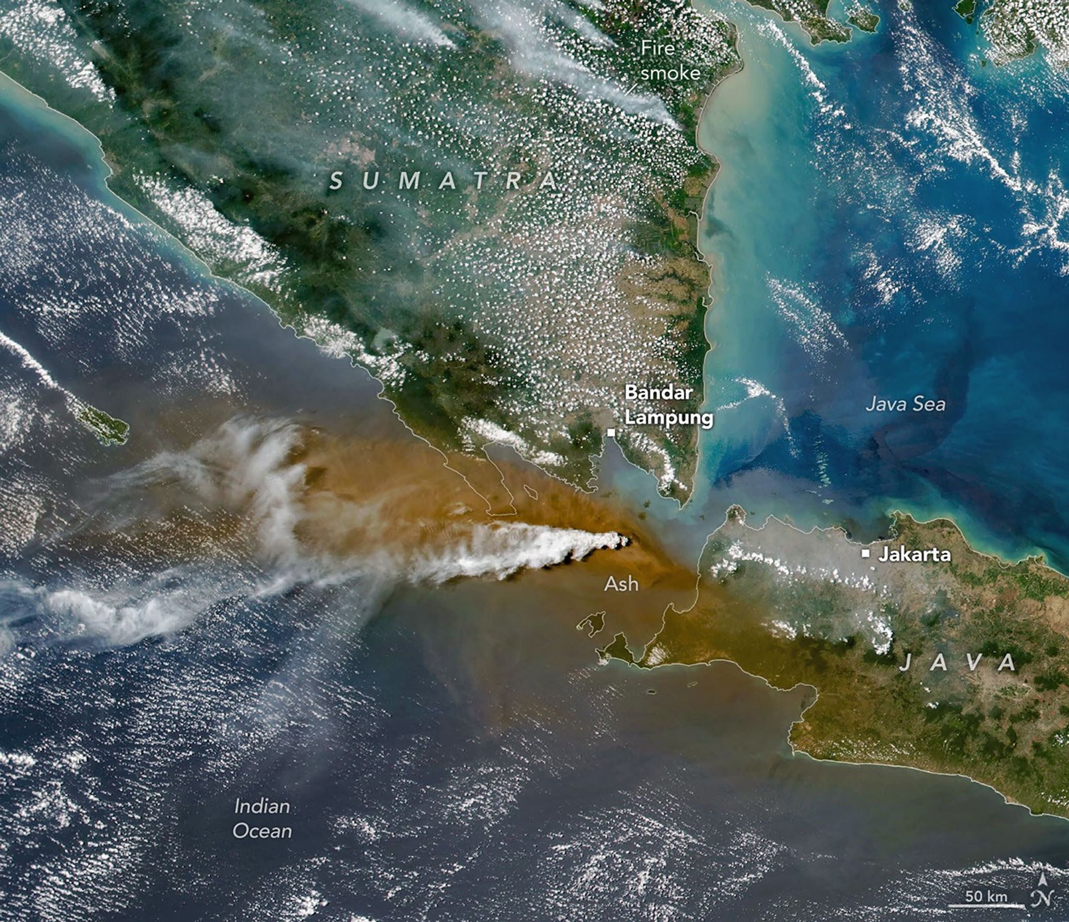 A white plume streams to the left over a larger tan ash cloud, both coming from a volcanic eruption between the Indonesian islands of Java and Sumatra.