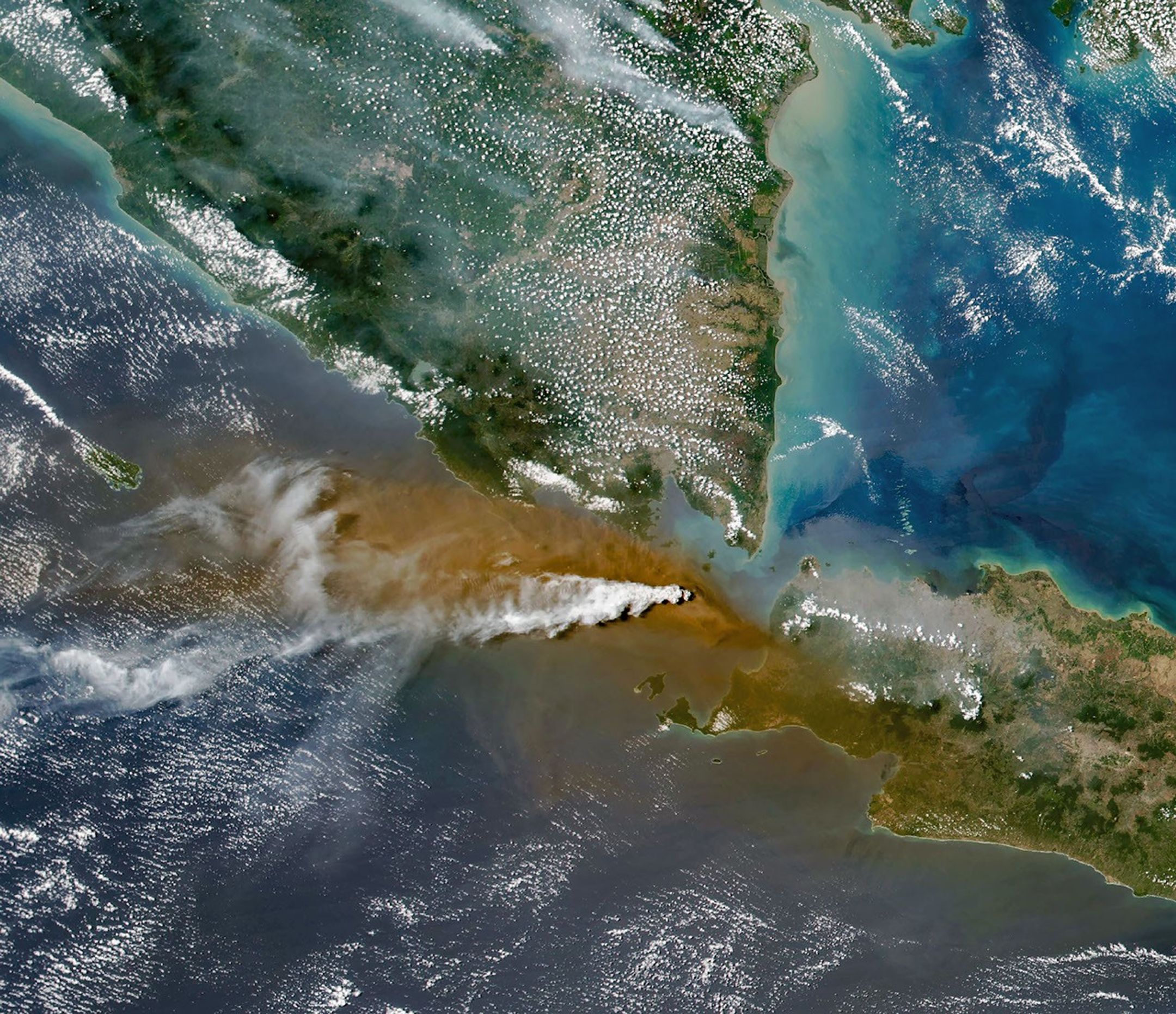 A white plume streams to the left over a larger tan ash cloud, both coming from a volcanic eruption between the Indonesian islands of Java and Sumatra.